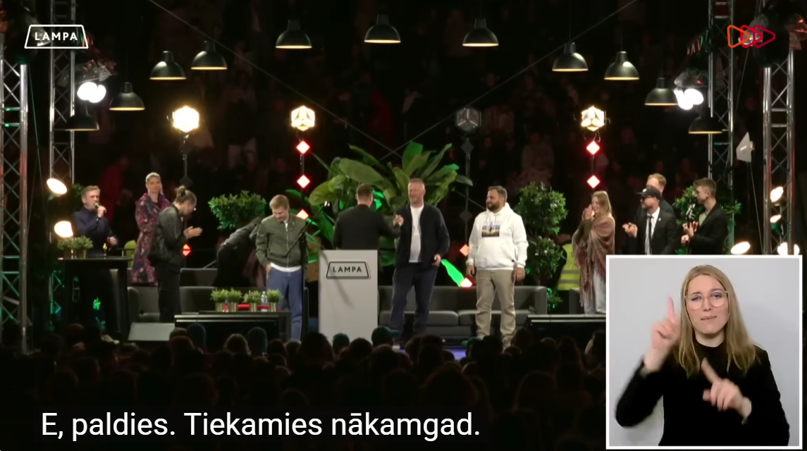 Ekrānā DOTS skatuve ar tiriem apakšā un ar surdotulkotāju
