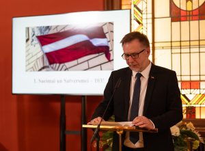 Ivars Balodis Saeimā lasot savu uzrunu. Ģērbies melnā uzvalkā un aiz muguras Latvijas karogs