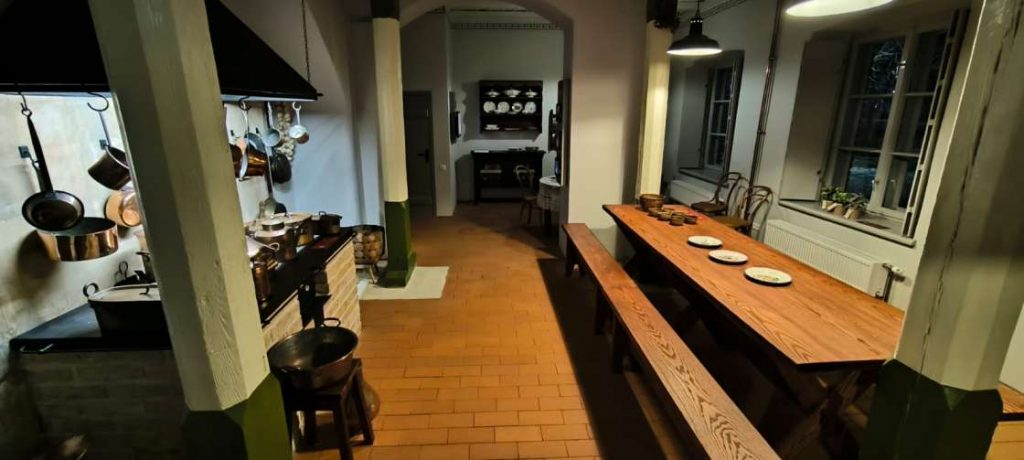 Ekspozīcija "Muižas virtuve" - brūns galds ar gariem brūniem soliem, gar sienu pannas, katli