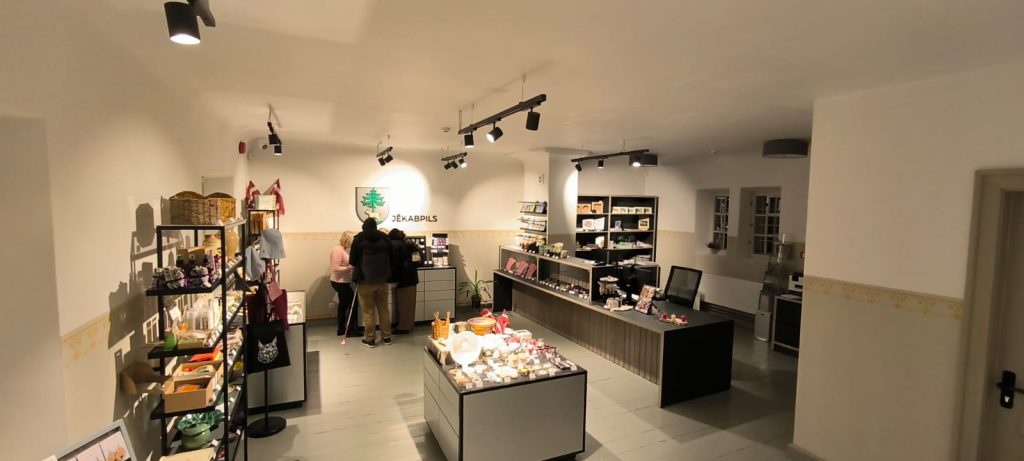 Apmeklētāju centra iekštelpa ar pilsētas ģērboni pie sienas, ir galdi un plaukti pilni ar suvenīriem