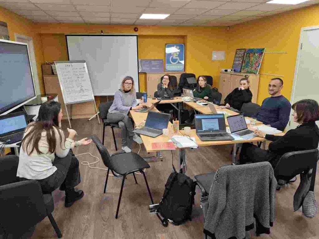Apeirons zālē vairāki cilvēki sēž pie galdiem, uz tiem ir laptopi, viena sieviete sēž atsevišķi. Ir 2 tāfeles