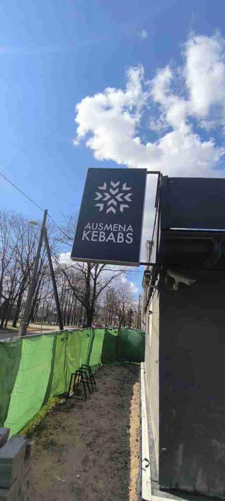 Pelēkas krāsas ēku apjož zaļas krāsas žogs, pie ēkas sienas novēro;sanas kamera un uzraksts Ausmeņu kebabs