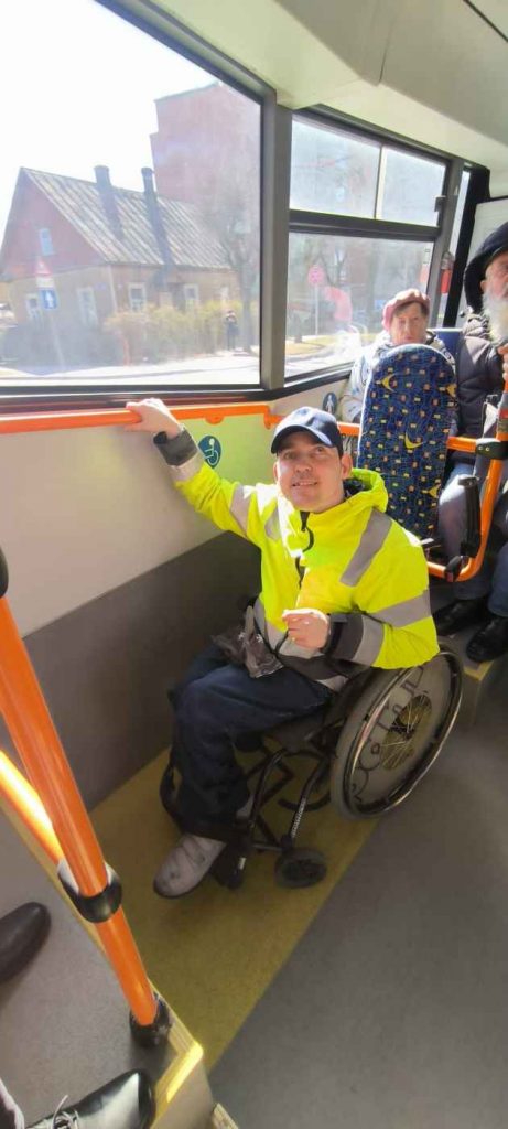 Voldemārs turas pie oranžas krāsas stieņa autobusā, aiz muguras zilas krāsas apmale