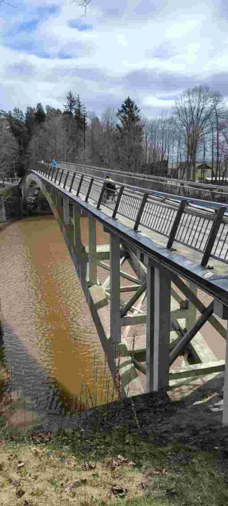 Trīs cilvēki šķērso Ogres upi pa tiltu, kuram ir augstas margas