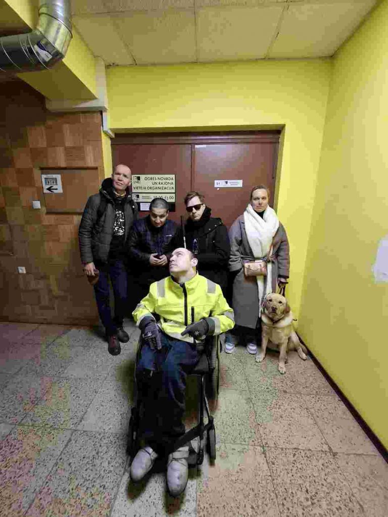 5 cilvēki pozē pie brūnas krās durvīm, sienas dzeltenzaļā krāsā