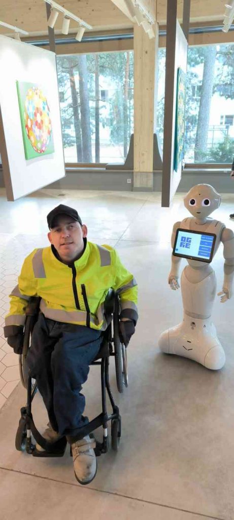 Voldemārs pozē ar baltas krāsas robotu, kuram priekšā ir displejs