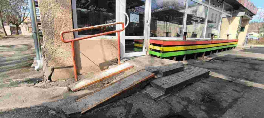 Uzbrauktuve pie kafejnīcas sadalīta divās daļās, augsta marga oranžā krāsā, pelēkas krāsas pakāpieni, durvis baltā krāsā