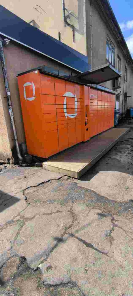 Pakomāts oranžā krāsā, priekšā viena pakāpiena dēļu grīda bez uzbrauktuves