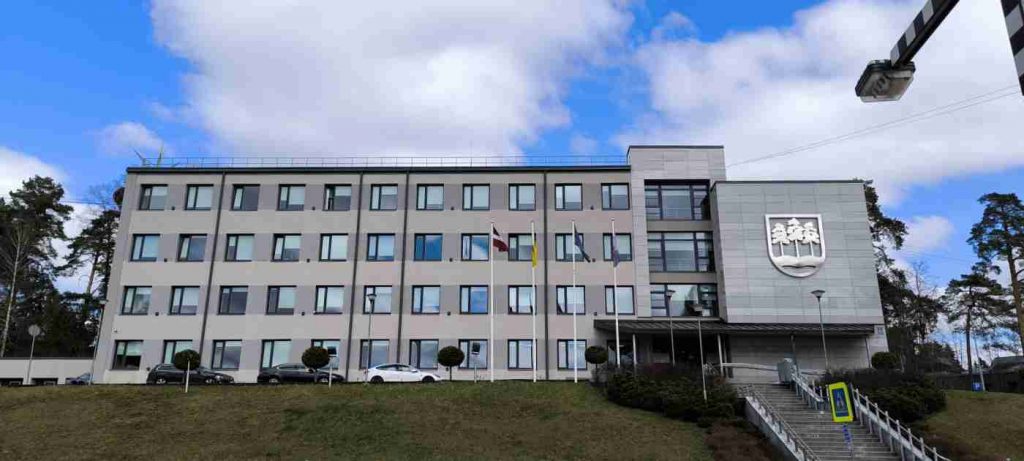 Fonā 4 stāvu pelēkas krāsas ēka ar Ogres logo, priekšā garas trepes