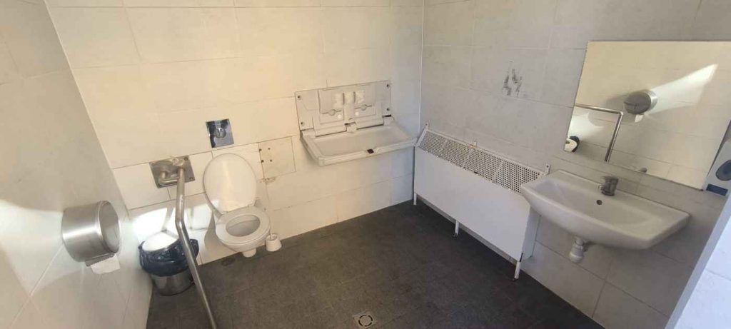 Tualetes iekšpuse baltā krāsā. Iekšā ir pods ar vienu rokturi, izlietne ar spoguli, bērnu pārtinamais galdiņš