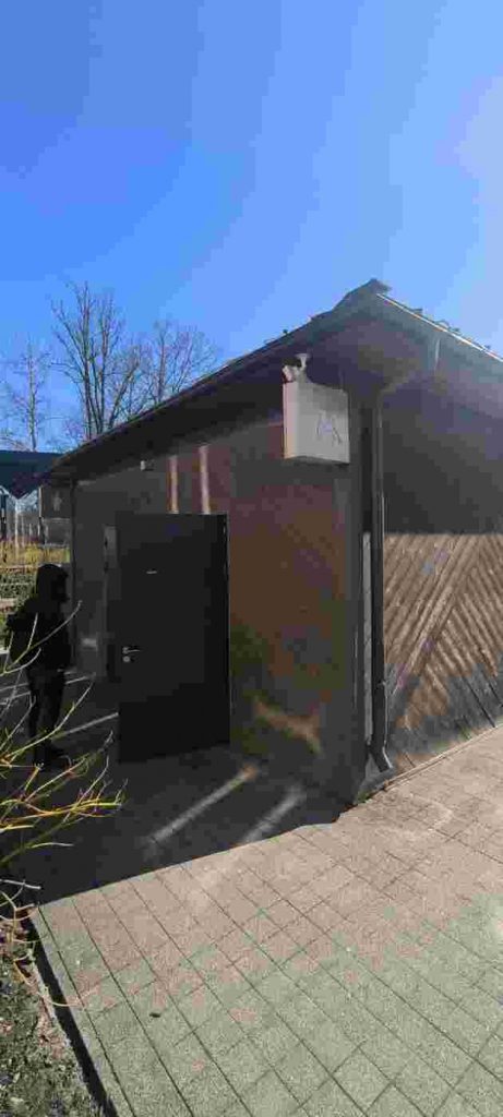 Tualetes ēka no koka brūnā krāsā, pie jumta cilvēku ratiņkrēslā zīme. Pie atvērtām durvīm cilvēks stāv.