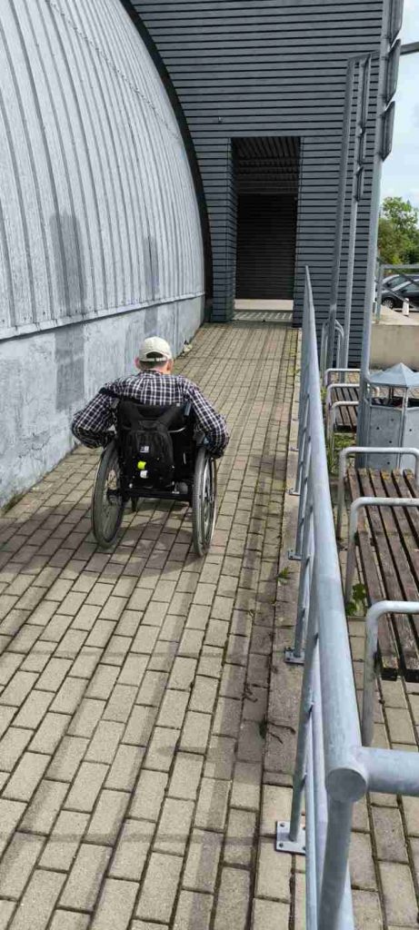 Cilvēks ratiņkrēslā pa labi bruģētu uzbrauktuvi dodas uz ieeju muzejā, vienā sānā margas, otrā angārs