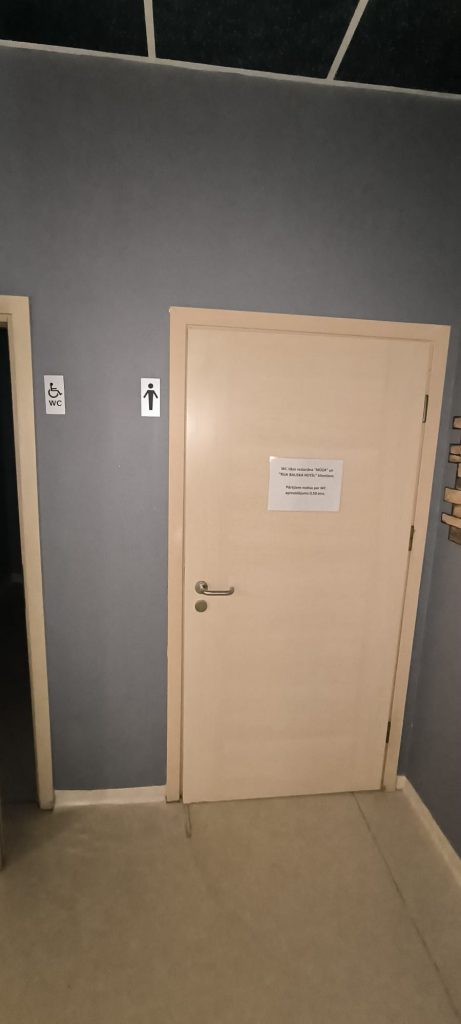 Gaiši brūnas krāsas tualetes durvis, pelēkas sienas, pie sienas diezgan augstu apzīmējums vīriešu un tualete cilvēkiem ratiņkrēslā