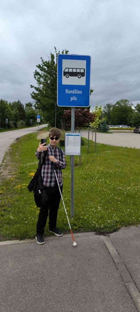 Cilvēks rūtainā kreklā, melnās biksēs ar balto spieķi taisa selfiju pie autobusa pieturas ar nosaukumu Rundāles pils