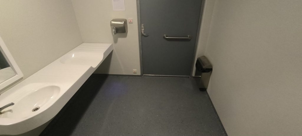 Tualetē balta izlietne, bērnu pārtinamais galdiņš, pie sienas roku žāvētājs, drošības poga, uz grīdas pie pelēkas krāsas izejas durvīm miskaste