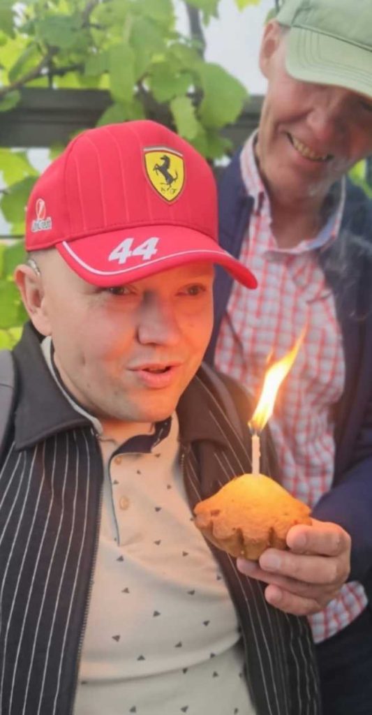 Cilvēks svītrainā žaketē, sarkanā beisbola cepurē pūš svecīti, kas iesprasuta keksiņā, aiz muguras cilvēks rūtainā kreklā, zilā jakā