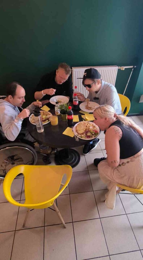 3 jaunieši un 1 jauniete ēd pusdienas pie melna galda dzeltenos krēslos