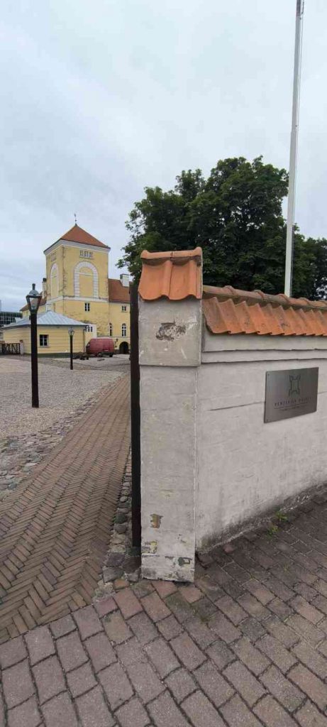 Dzeltenā krāsā Livonijas ordeņa pils, bruģēts celiņš uz pili, priekšā mūra žogs ar dakstiņu jumtiņu