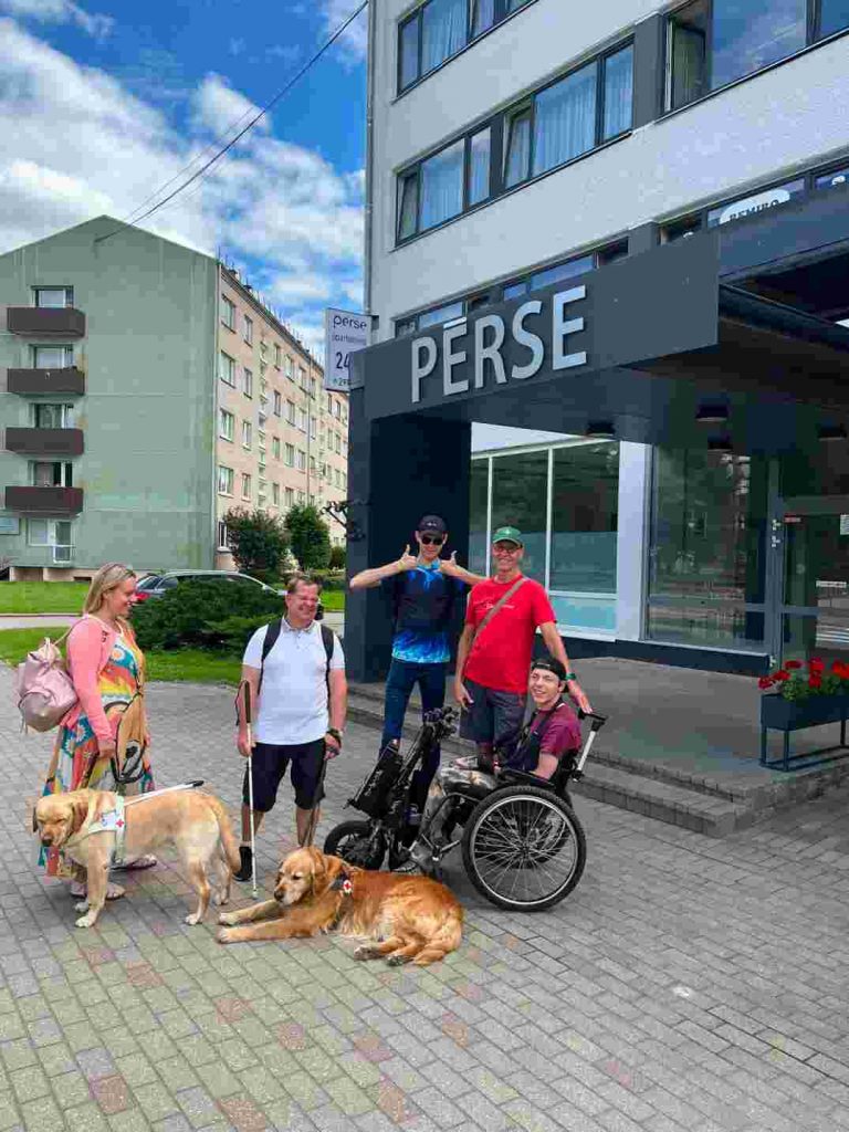 5 cilvēki ar suņiem-pavadoņiem pie 4 stāvu ēkas ar uzrakstu Pērse