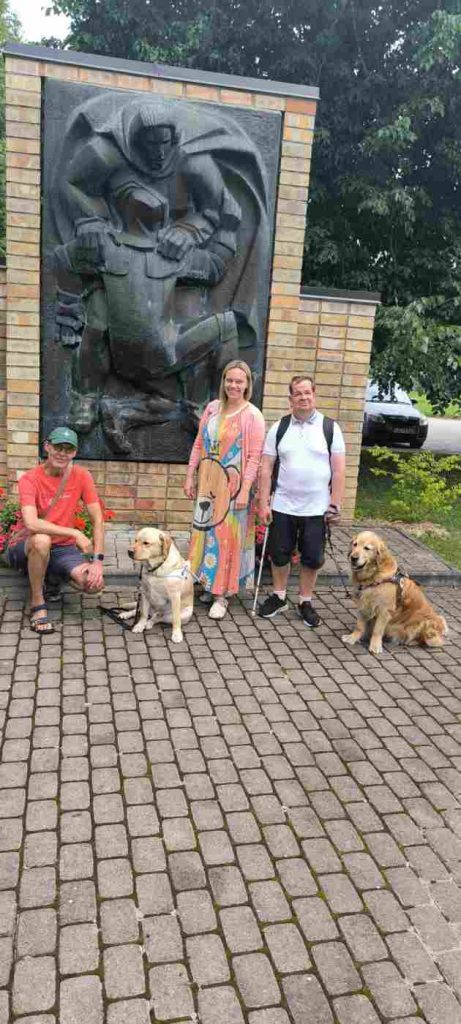 3 cilvēki un suņi-pavadoņi pie pieminekļa, kurš ir novietots uz dzeltenas krāsas ķieģeļiem