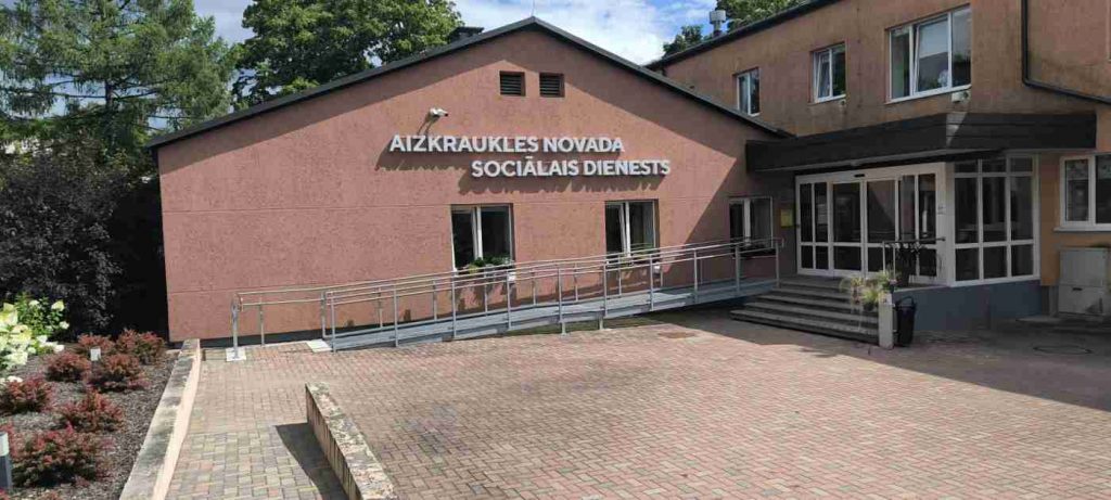 Divstāvu brūnas krāsas ēka ar uzrakstu Aizkraukles novada sociālais dienests. Ēkas priekšā uzbrauktuve ar margām