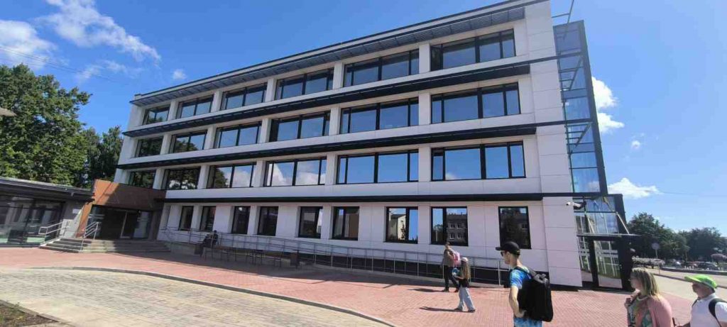 4 stāvu ēka gaišās krāsās, gar ēku ir uzbrauktuve ar margām. Ēkas priekšā 5 cilvēki