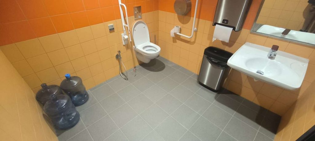 Tualetē pelēka flīžu grīda, uz sienas dzeltenīgas un oranžas flīzes, ir pods, rokturi, izlietne, spogulis, drošības aukla, miskaste,papīra dvieši. Stūrī 3 izlietotas Venden pudeles zilā krāsā