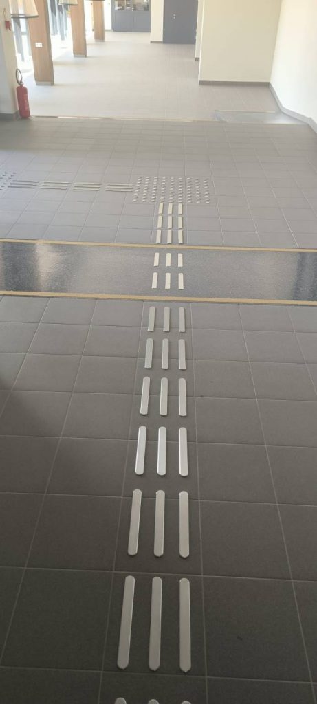 Ēkas iekšpusē uz pelēkas grīdas flīzēm dzelzs vadlīnijas