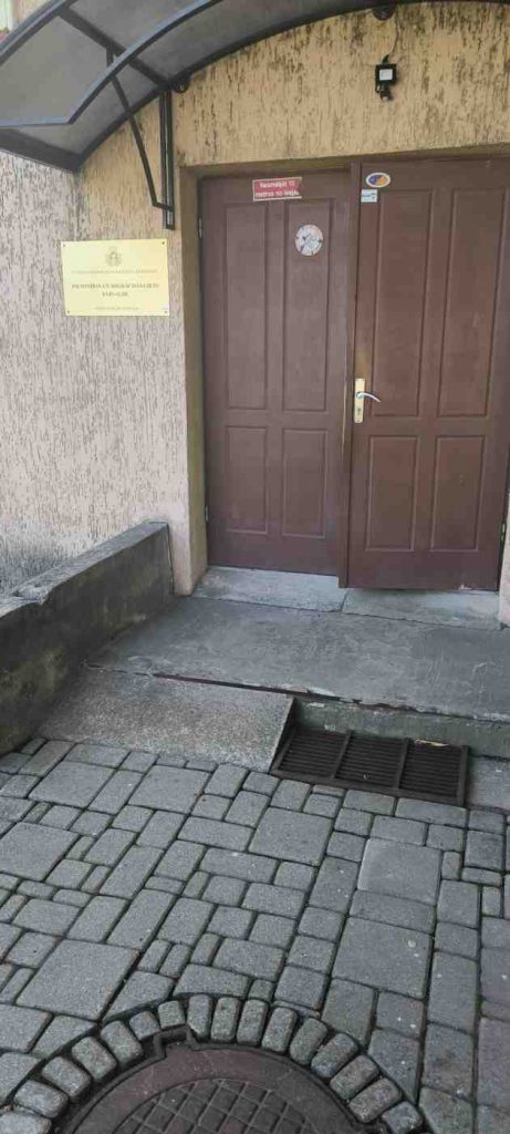 Ēka, kurai ir brūnas durvis. Ēkas priekšā ir diezgan stāva uzbrauktuve
